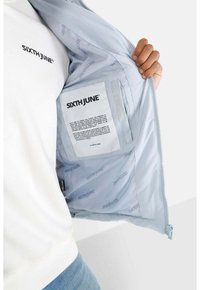 Giacca azzurro chiaro con chiusura a zip, foderato internamente con un marchio che presenta testo ripetuto, e una tasca per etichetta sul lato sinistro dell'interno.