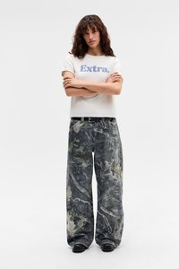 Weißes Baumwoll-T-Shirt mit "Extra." in blauer Schrift, kombiniert mit locker sitzenden, gemusterten grau-grünen Hosen und schwarzem Schuhwerk.