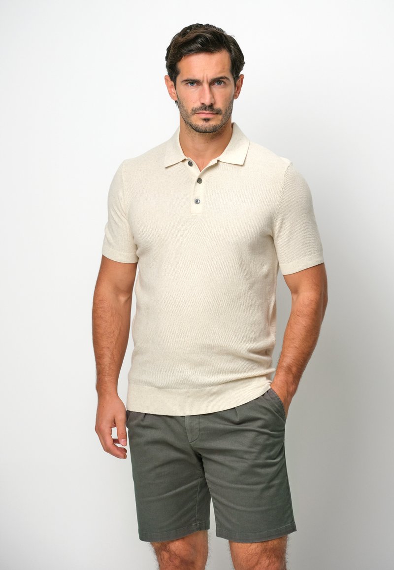 Beige Polo-Shirt mit kurzem Arm und strukturiertem Muster, drei Knöpfen und geripptem Saum. Getragen mit dunkelgrünen Shorts.