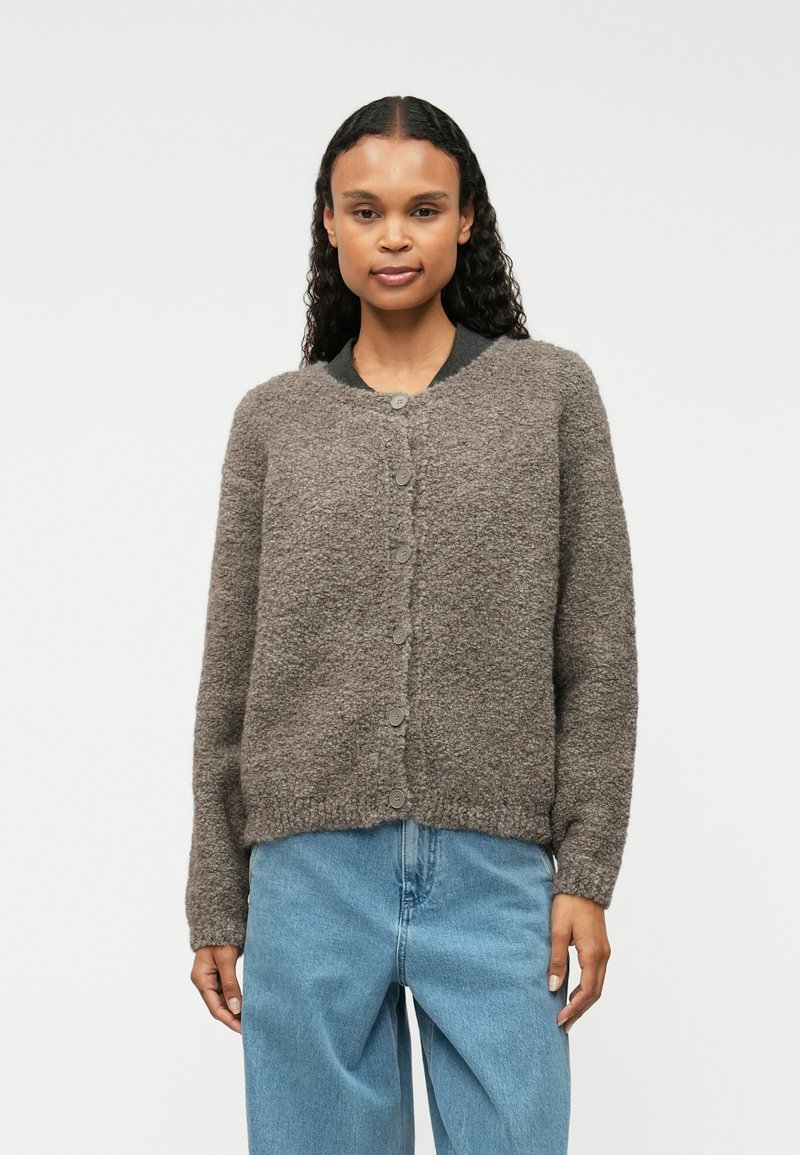 Sivý, štruktúrovaný kardigán s okrúhlym výstrihom a zapínaním na gombíky, kombinovaný so svetlo modrými džínami. Mäkka tkanina a voľný strih.