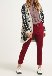 Strickjacke mit schwarz-weißem Leopardenmuster, über einer grauen Bluse mit Halsband getragen, kombiniert mit roten Hosen und weißen Schuhen.