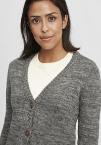 Grau gestrickte Strickjacke mit V-Ausschnitt, ausgestattet mit einem strukturierten Muster und zwei braunen Knöpfen an der Vorderseite. Über einem hellen Hemd getragen.