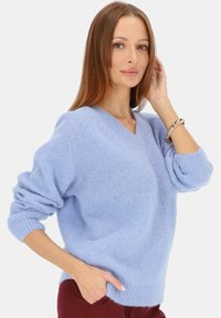 Jasnoniebieski, miękki sweter dziany z dekoltem w serek i podwiniętymi mankietami. Materiał wygląda na puszysty, oferując luźny krój. Noszony z burgundowymi spodniami.