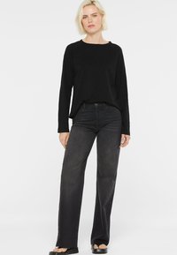 Blonde Frau, die ein schwarzes Langarmshirt, hochgeschnittene schwarze weite Jeans und schwarze Plateausandalen trägt.