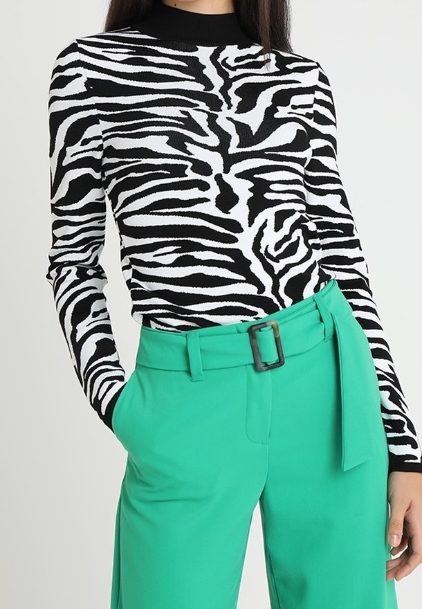 Kobieta w czarno-białej bluzce w zebry z długimi rękawami, włożonej w zielone, wysokie spodnie z paskiem, z rękami w kieszeniach.