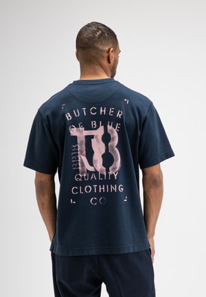Man met zijn rug naar de camera, draagt een marineblauw T-shirt met grote roze grafische tekst "Butcher of Blue T8 Quality Clothing Co" op de achterkant.