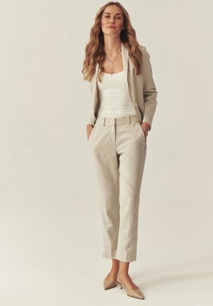 Frau mit langem, welligem Haar, die einen beigen Anzug, ein weißes Oberteil und beige Absatzschuhe trägt, mit den Händen in den Taschen vor einem schlichten Hintergrund steht.