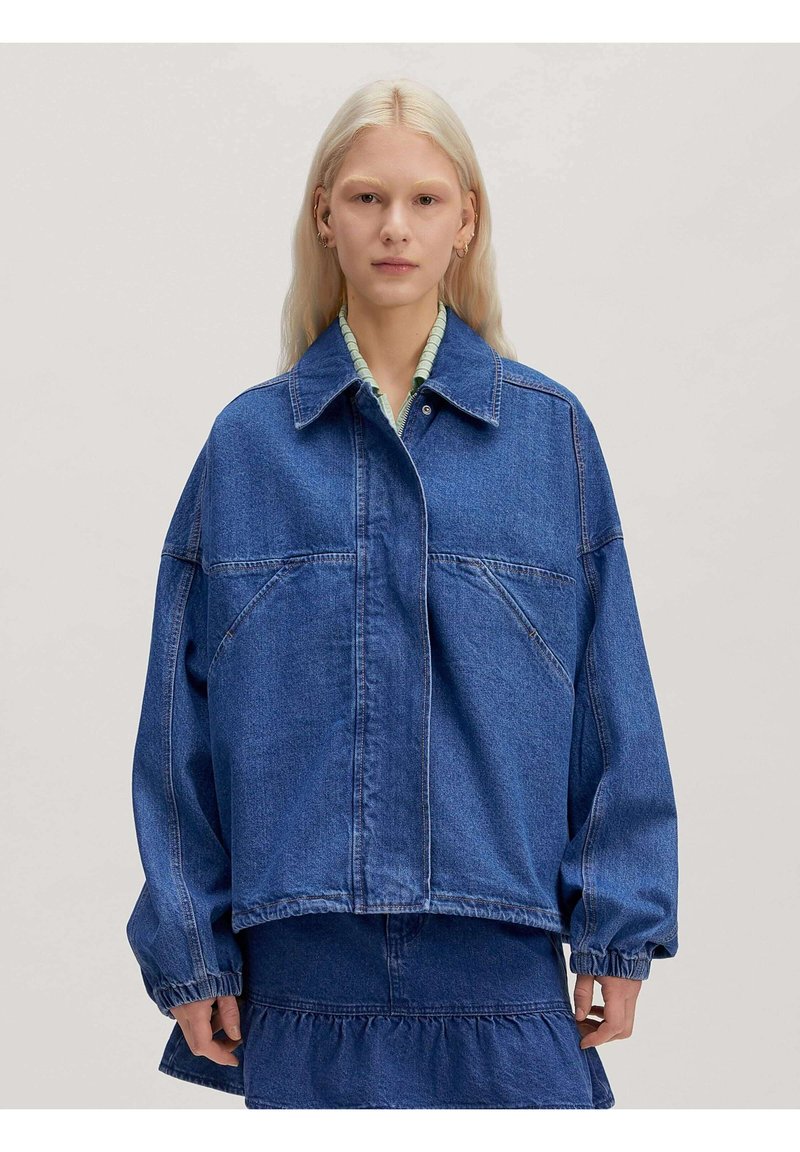Blondine vrouw die een oversized blauwe spijkerjas en bijpassende rok draagt, poserend tegen een effen lichtgrijze achtergrond.
