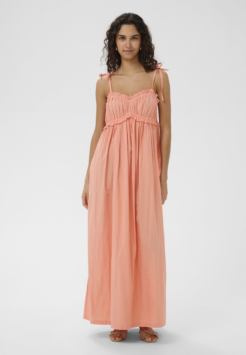 Femme aux cheveux bouclés portant une robe maxi sans manches de couleur pêche avec des bretelles nouées aux épaules et des sandales, debout devant un fond uni.