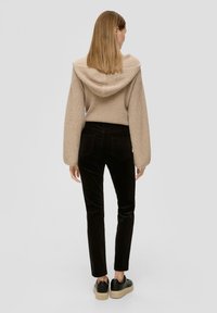 Femme avec des cheveux droits châtain clair portant un hoodie en tricot beige, un pantalon noir et des baskets compensées noires, se tenant de dos sur un fond gris.