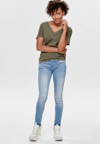 Olivově zelené tričko s výstřihem do V, volnější střih, spárováno se světle modrými skinny džíny a bílými teniskami, vnitřní prostředí, neutrální pozadí.