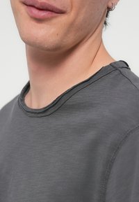 Close-up of lower face and neck wearing dark grey crew neck shirt with raw-edge hem and small hoop earrings on left ear.