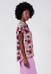 Blusa floreale a maniche corte in rosa, con grandi fiori viola e gialli e colletto. Tessuto morbido, con fessure laterali per facilità.