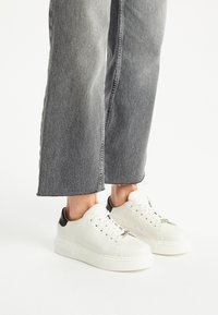 Białe sneakersy z czarnym akcentem na pięcie, z perforacjami, płaskim sznurowaniem i masywną podeszwą; noszone z szerokimi szarymi dżinsami.