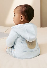 Felpa con cappuccio azzurro chiaro lavorata a maglia con un disegno di orso sul retro. Presenta una texture morbida e un cappuccio con coulisse. Sono visibili pantaloncini a righe.