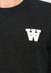 Svart strikket genser med ribbestruktur og en fremtredende hvit "W" logo på brystet. Rund halslinning og tettsittende design.