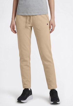 Pantalones jogger beige con cordón y bolsillos delanteros, combinados con zapatillas negras y blancas y una camiseta gris.