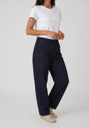 Mujer con camiseta blanca de manga corta, pantalones anchos azul oscuro y zapatillas beige, de pie contra un fondo liso claro.