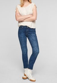 Top acanalado de color beige claro con detalles de lazo en los hombros, combinado con vaqueros skinnies azul oscuro con estampado y zapatillas altas blancas.