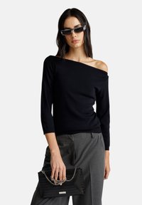 Top de punto negro sin hombros con mangas 3/4, combinado con pantalones grises de corte recto y un bolso de mano negro texturizado con un acento de cadena.
