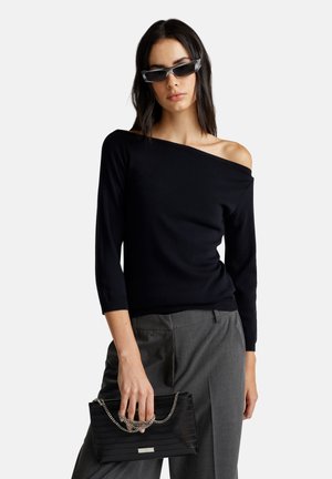 Haut en tricot noir à épaules dénudées avec des manches 3/4, associé à un pantalon gris ajusté et un sac à main texturé noir avec un accent en chaîne.