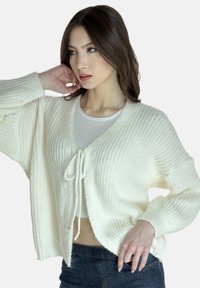 Weißer Strickcardigan mit V-Ausschnitt, lockerer Passform und vorderem Bindeband. Verfügt über ribbelige Textur und weite Ärmel. Trägt man über einem hellen Oberteil.