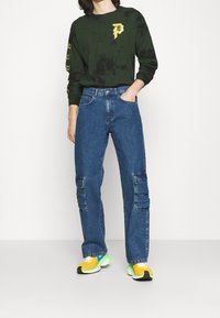 Dark green long-sleeve shirt with yellow graphic, paired with blue cargo jeans featuring side pockets and yellow-and-green sneakers.