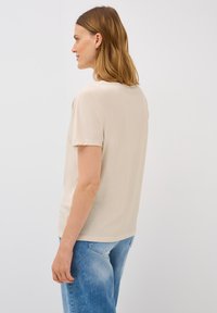 Beige t-shirt med kort ärm i mjukt material med rund halsringning, avslappnad passform och en liten logolapp vid fållen. Matchas med ljusblå jeans.