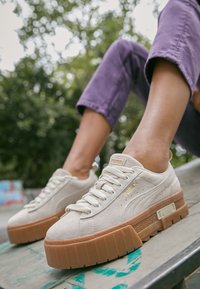 Baskets en daim beige avec une semelle épaisse de couleur tan, présentant des accents de logo dorés et un laçage traditionnel, sur un fond extérieur flou.