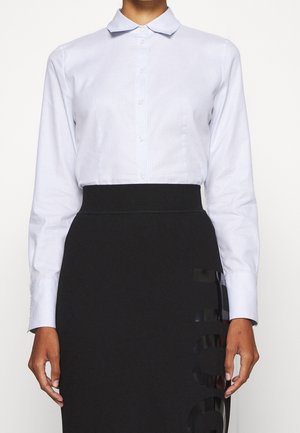 Persoon die een lichtblauw overhemd draagt, ingestopt in een hoge taille zwarte rok met glanzende zwarte letters aan de zijkant.