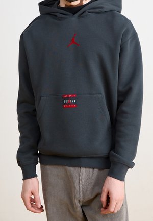 Osoba nosząca czarną bluzę z kapturem Jordan z czerwonym logo Jumpman i naszywką "Authentic Jordan Brand" na przedniej kieszeni, w zestawieniu ze sztruksowymi spodniami.