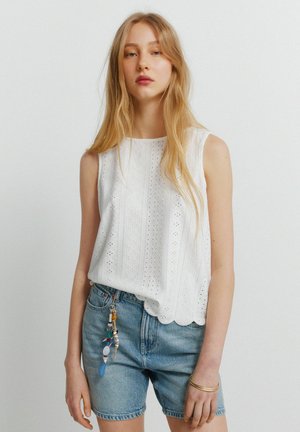 Ung kvinde med langt blondt hår iført hvid ærmeløs bluse med hulmønster og blå denimshorts med nøglekæde med perler.