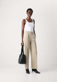Vrouw in een witte tanktop en beige wijde broek, met een zwarte handtas en zwarte spitse enkellaarsjes voor een witte achtergrond.