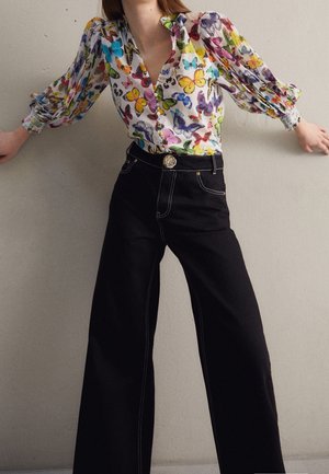 Femme portant un chemisier coloré à imprimé papillons avec des manches bouffantes et un pantalon large noir avec des surpiqûres blanches et un bouton décoratif.