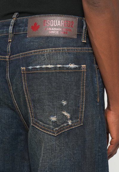 Jeans de mezclilla azul oscuro con un acabado desgastado. Presentan un parche de cuero marrón con letras rojas y costuras visibles en los bolsillos.