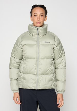 Persona con chaqueta acolchada verde clara con logo de Columbia, de pie contra un fondo liso, con las manos relajadas a los lados.