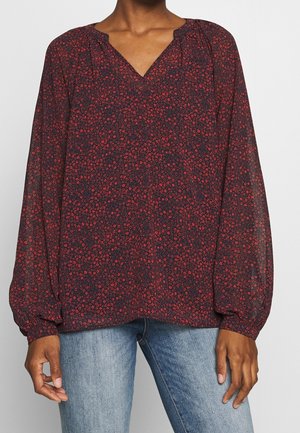 Kobieta ubrana w ciemną bluzkę z czerwonym wzorem kwiatowym, długie bufiaste rękawy, dekolt w serek, w połączeniu z jasnoniebieskimi dżinsami.