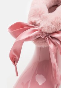 Botte cheville en vernis rose avec un col en fausse fourrure rose duveteuse et un ruban en satin assorti noué en haut. Surface lisse et brillante avec une texture douce.