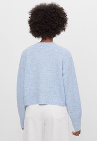 Person med lockigt hår har på sig en ljusblå fluffig tröja och vita byxor, står vänd bortåt mot en enkel vit bakgrund.