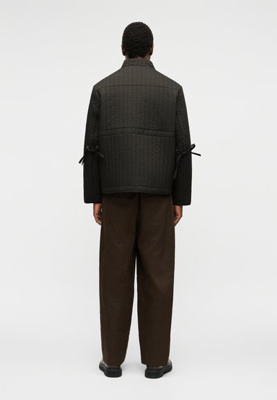 Personne de dos portant une veste matelassée noire avec des détails noués aux manches et un pantalon ample marron foncé sur un fond clair uni.