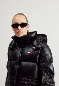 Chaqueta de plumas negra con un cuello alto, textura brillante, cierre de cremallera y parche de logo bordado. Combinada con elegantes gafas de sol negras.