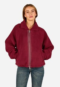 Burgundi värvi fliisjakki, millel on lai krae ja täispikk tõmblukk, üle mõõdu suurte varrukatega ning tekstureeritud pind.