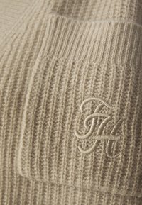 Beige stickad tröja med ribbad struktur och en framficka. En broderad logotyp är placerad på den nedre högra sidan.
