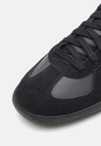 Svart sneakerspets med mocka och släta läderpaneler, svarta snören och strukturerad svart gummisula på vit bakgrund.