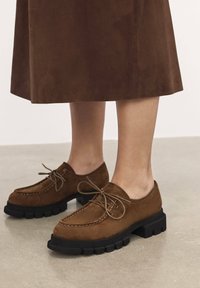 Pantofi din nubuc maro, cu o talpă groasă neagră, av�ând un design cu șireturi și detalii de cusătură pe partea superioară.