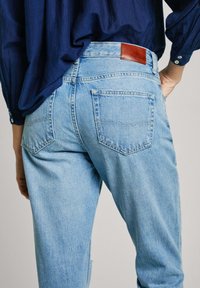 Šviesiai mėlynos džinsinės kelnės su aukštu liemeniu, dviem užpakalinėmis kišenėmis ir rudos odos etikete ant liemens. Minkšta tekstūra ir laisvas stilius.