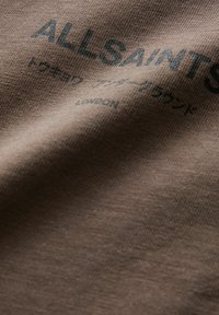 Ruda medvilnė su lygiu paviršiumi, dekoruota tamsiai žaliais spausdintais tekstais, įskaitant prekės ženklą "ALLSAINTS" ir kitą informaciją.