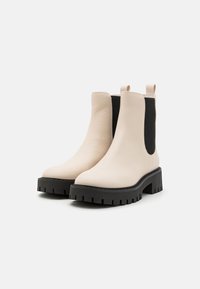 Beżowe skórzane botki za kostkę z masywną czarną gumową podeszwą. Zawierają elastyczne panele boczne oraz uchwyty do łatwego zakładania. Teksturowane wykończenie.