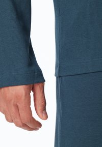 Mano izquierda relajada al lado del cuerpo, vistiendo una camiseta de manga larga azul con canalé y pantalones azules a juego, contra un fondo blanco.