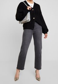 Svart stickad cardigan med stora knappar, grå rak benjeans och högklackade skor i orm-tryck. Modellen håller en flerfärgad handväska med detaljer.